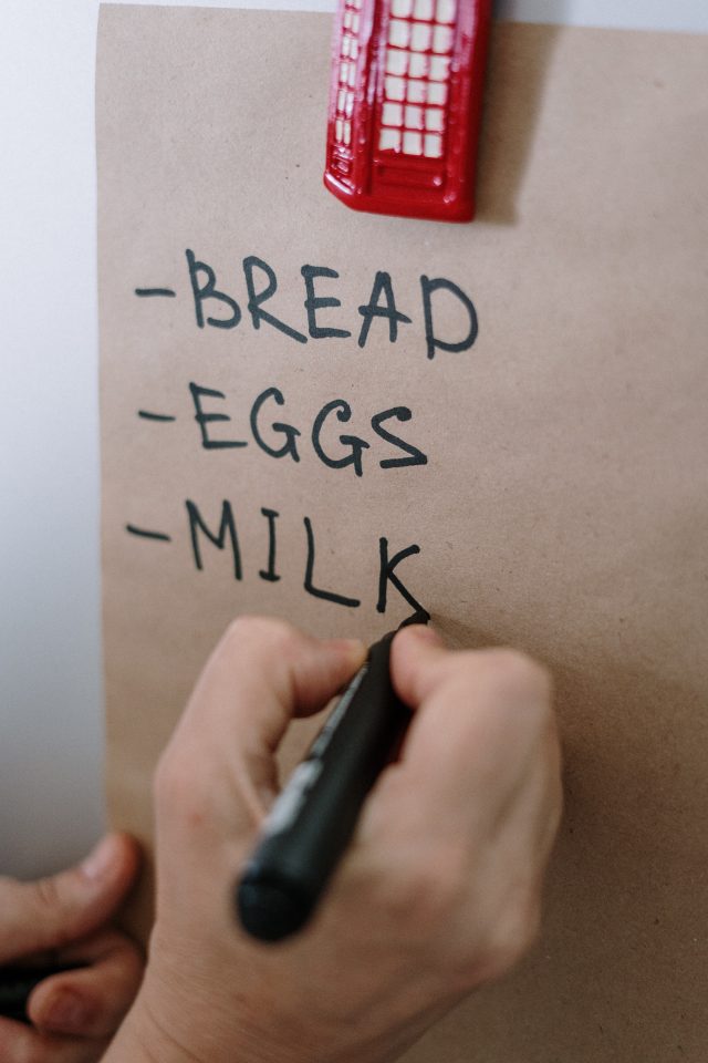a shopping list attached to a fridge with someone writing on "bread, eggs and milk"