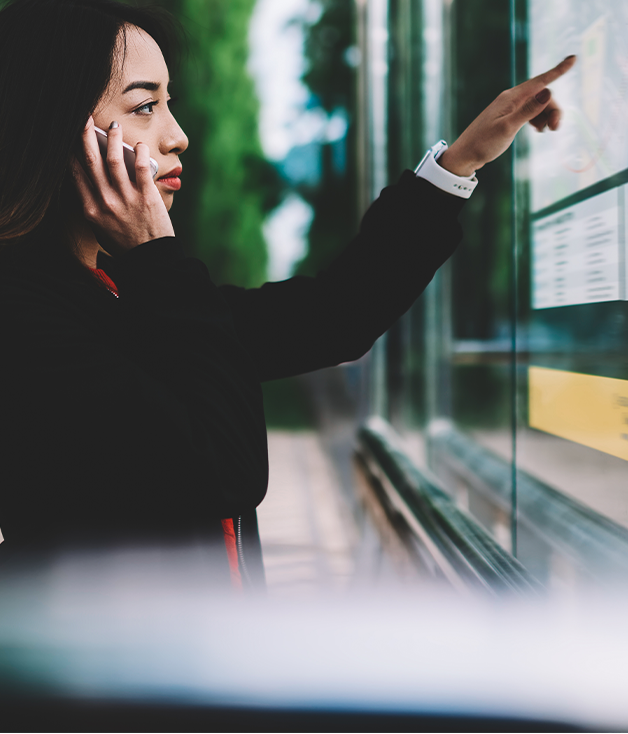 a young girl stood with her phone to her ear, whilst trying to read a bus timetable