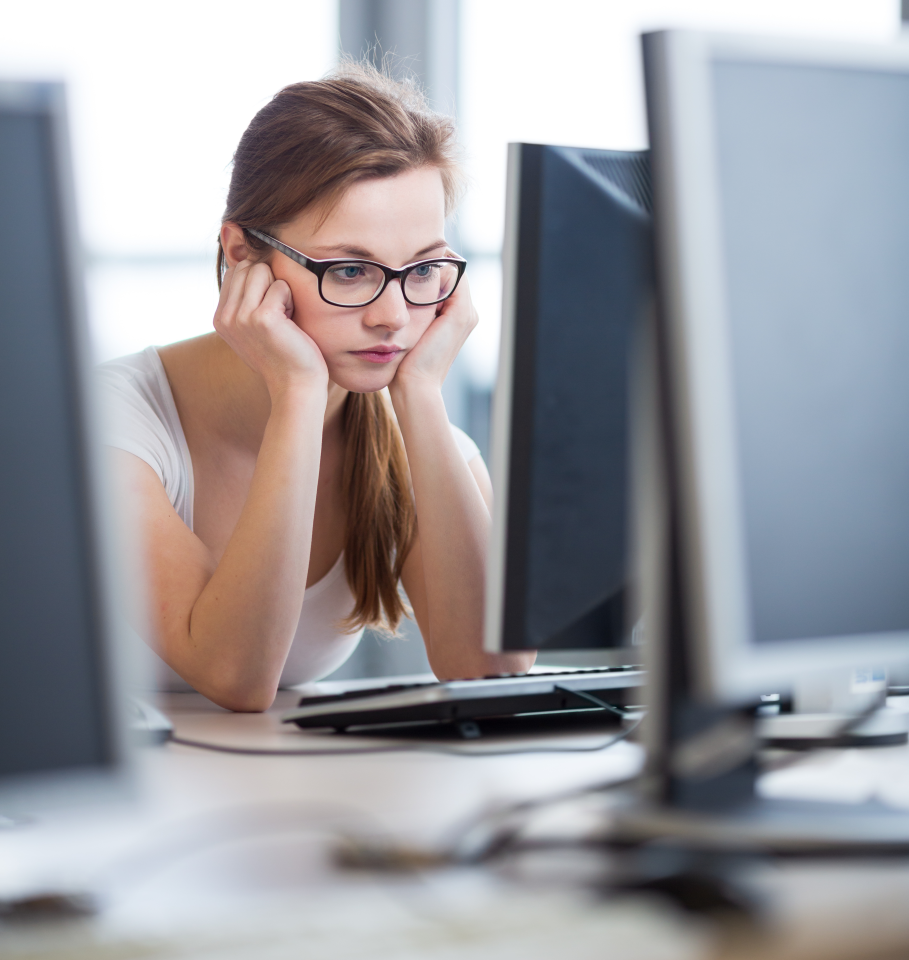 a young female office worker is sat in front of their desktop computer reading a document. They have their head in their hands whilst looking at the screen, looking fed up and confused with what they are reading.