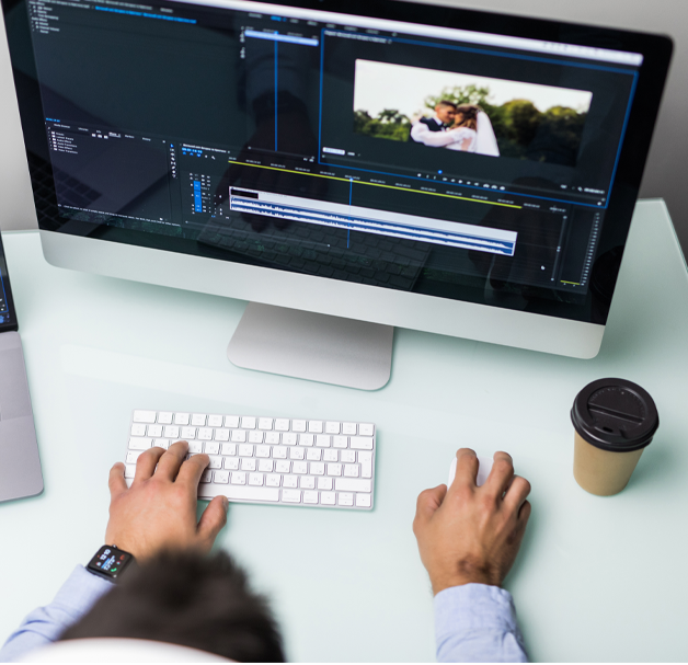 a desktop computer on a pale green desk has Adobe Premiere Pro on the screen. Taken from above, a man is sat editing a video and adding captions using a Mac.