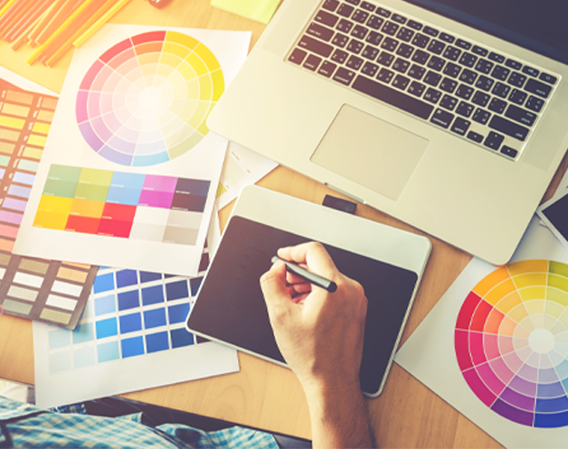 A graphic designer is sat at a desk in front of a laptop and using a drawing tablet. Surrounding him are a series a colour wheel charts.