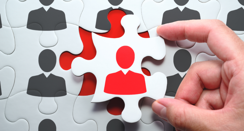 A close-up of a hand holding a puzzle piece that highlights a single red figure standing out from several grey figures.